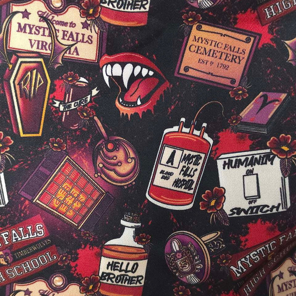 A close-up of a black fabric printed with a repeating gothic horror pattern featuring tombstones, vampire fangs, a potion bottle, flowers, and text such as 'Mystic Falls Cemetery' and 'Hello Brother'.